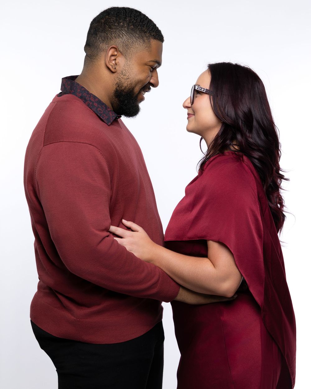 A couple in burgundy outfits embraces and laughs together in a series of candid poses against a white backdrop.