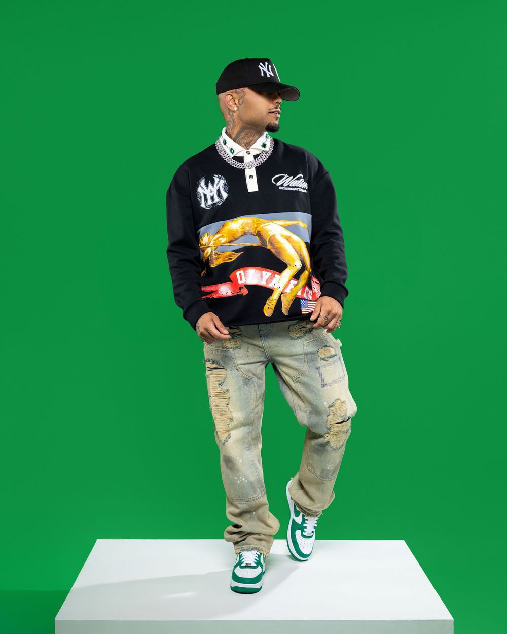 Person in black jacket and distressed jeans poses on white platform against green background showing urban streetwear style.
