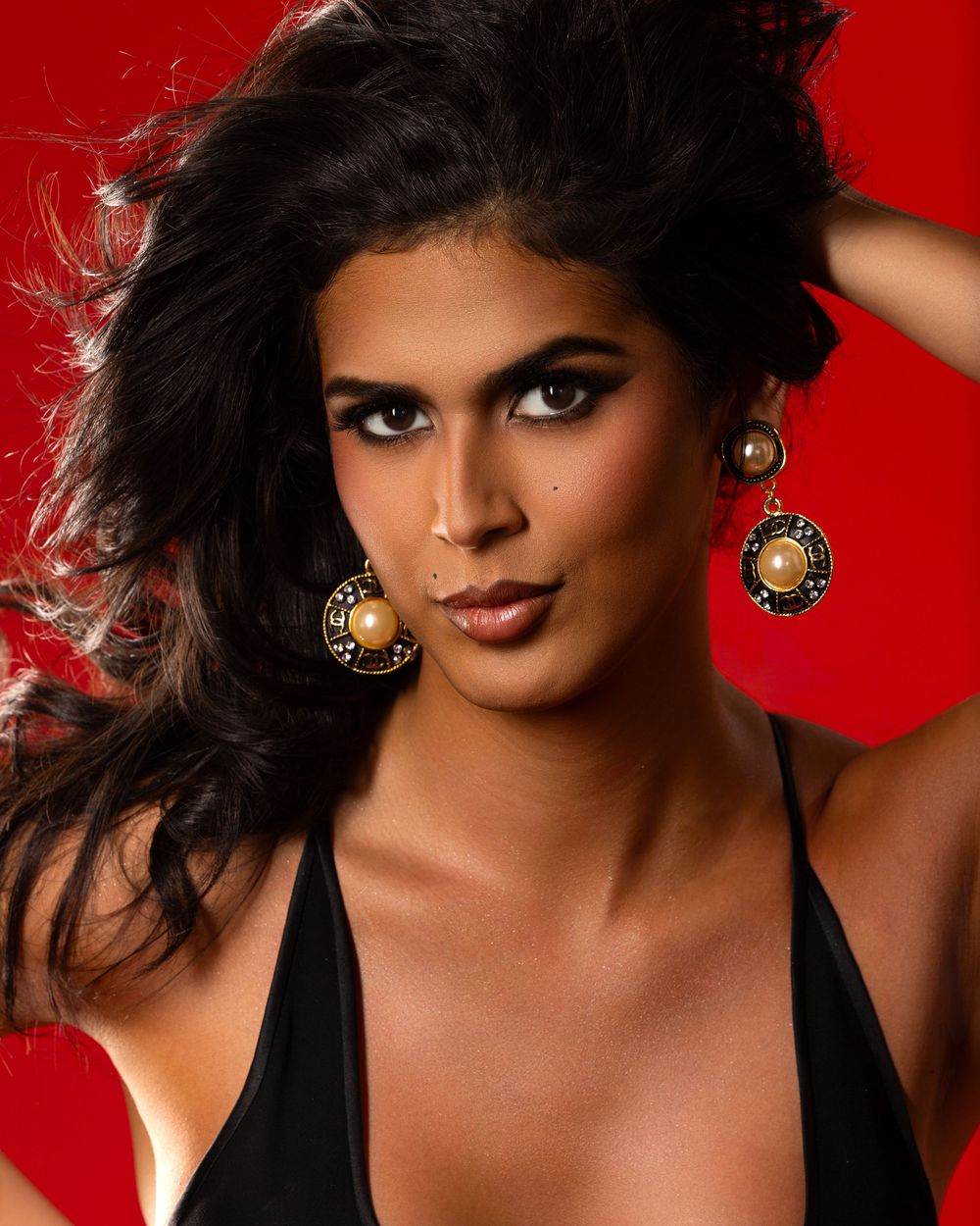 Portrait against bold red background showcasing glamorous makeup, dark wavy hair and ornate earrings in dramatic style.