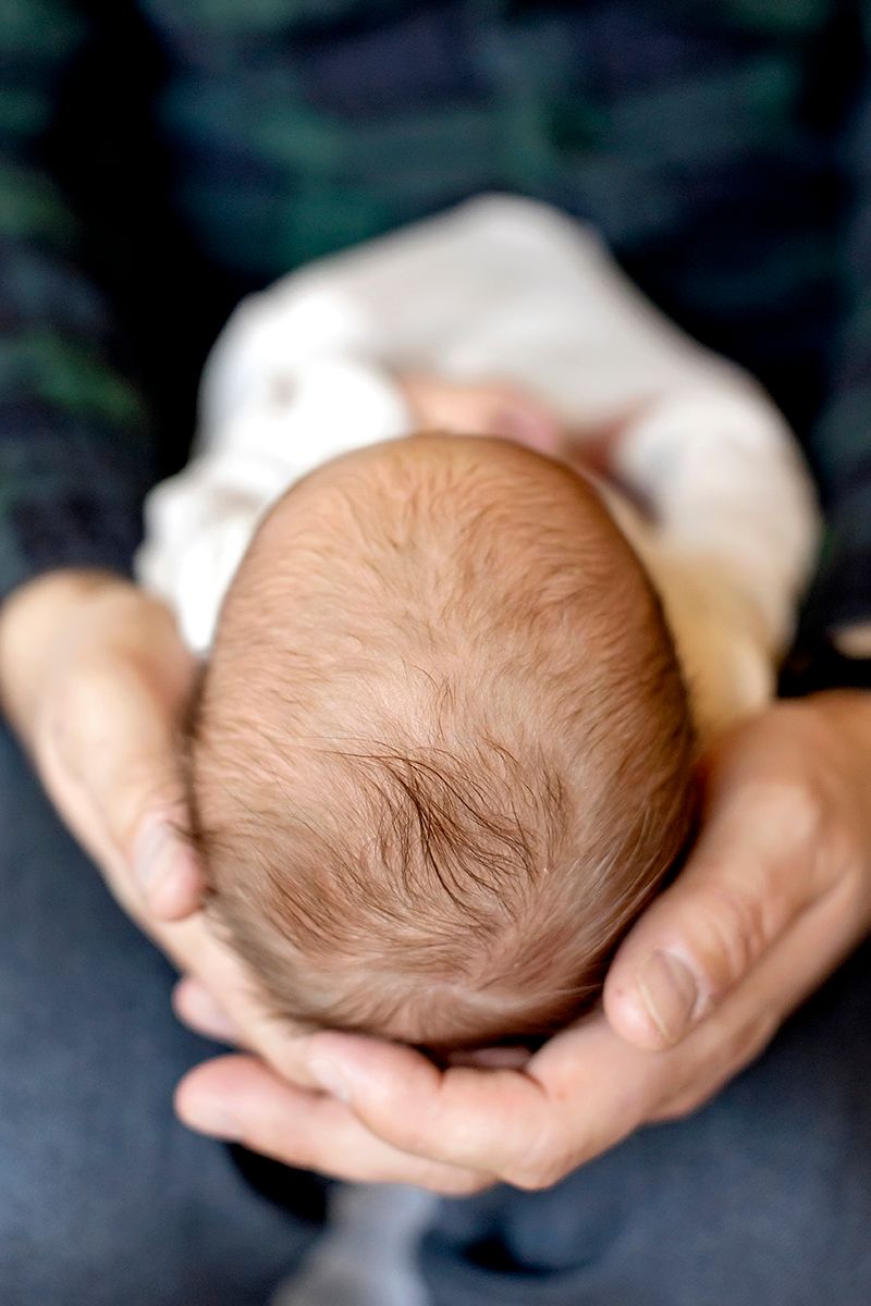 Gentle hands cradle a newborn baby's head showing soft brown hair against a dark background.
