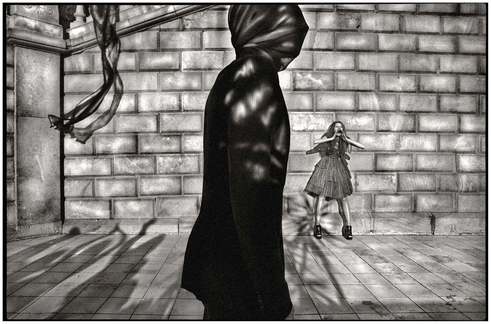 Black and white photo of cloaked figure and girl in striped dress against stone wall with dramatic shadows.