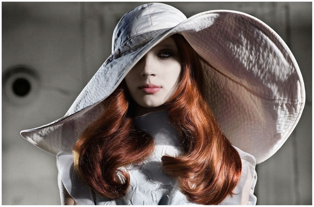 A woman with long red hair wearing a wide-brim white hat and white outfit poses elegantly.