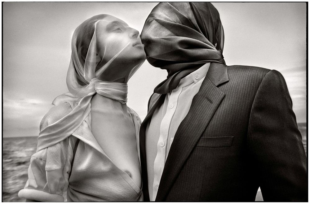 Black and white photo of a couple kissing with scarves blowing over their faces in the wind outdoors.