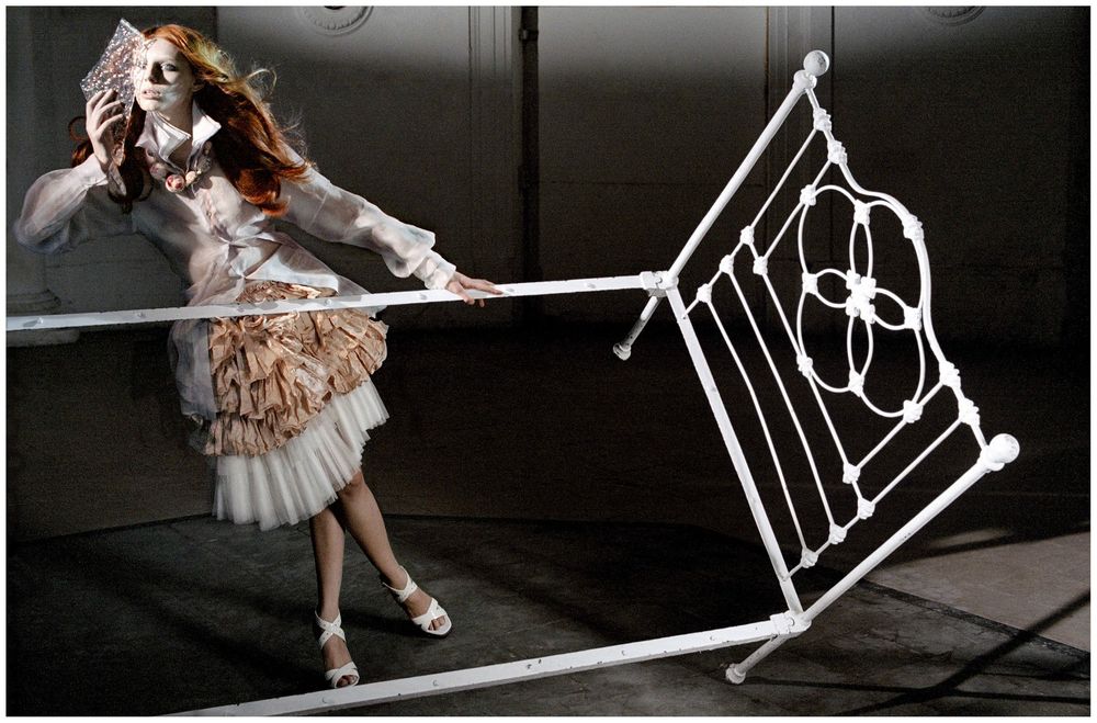 Fashion model in elegant dress pulling an ornate white metal bed frame in a dramatic studio setting.