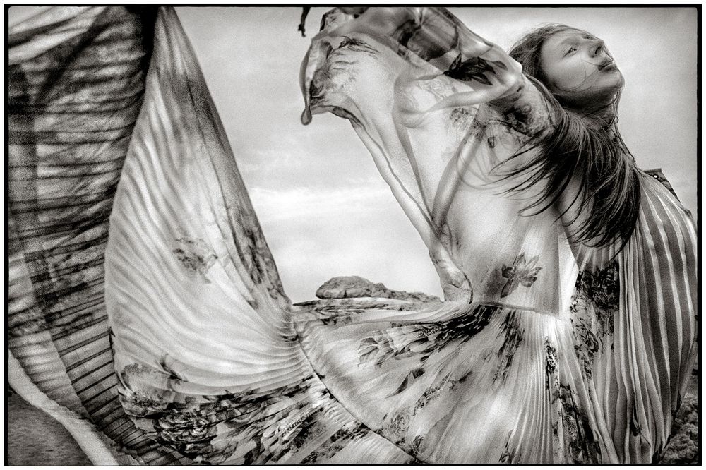 Black and white photo of a woman in a flowing patterned dress, fabric billowing dramatically in the wind outdoors.