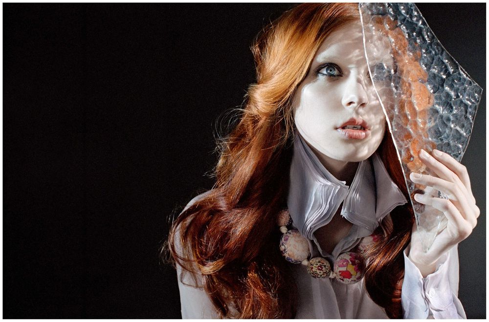 Red-haired woman with pale skin holds sheer fabric near her face against a dark background.
