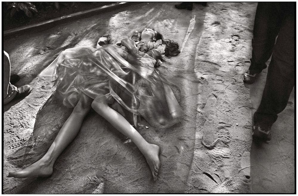 Black and white photo of a body covered in plastic sheeting lying on muddy ground, bystanders visible nearby.