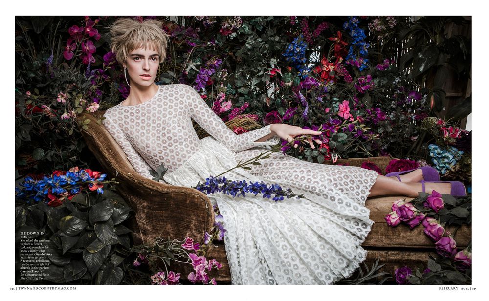 A person in a white lace dress reclines on a wooden bench surrounded by vibrant pink and blue flowers.
