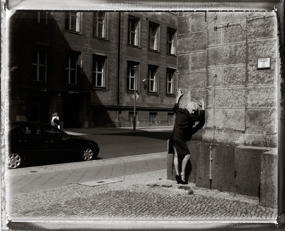 A black and white panoramic sequence shows a person walking past stone buildings on a cobblestone street.