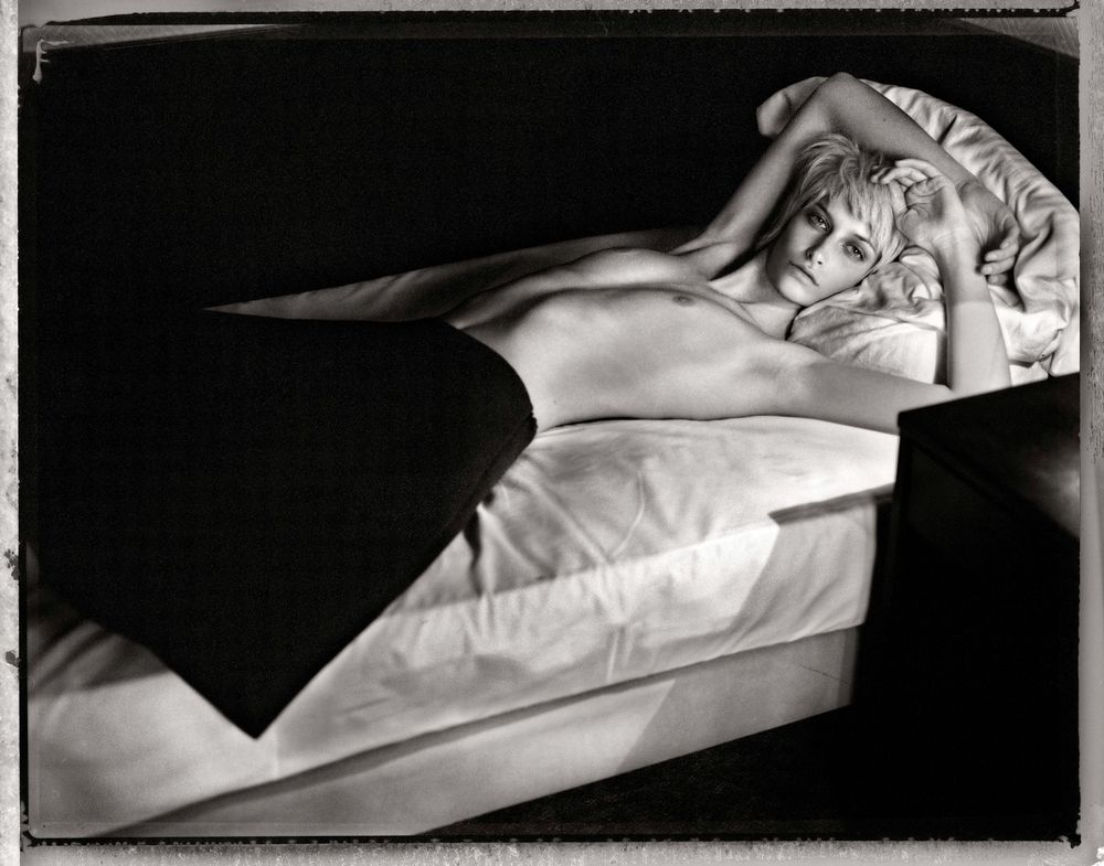 Black and white photograph shows a series of rumpled white bed linens and pillows arranged horizontally.