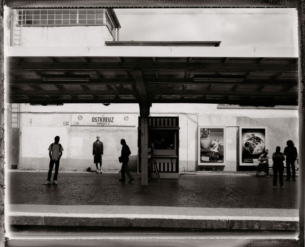 A panoramic black and white photograph showing multiple silhouettes of people waiting at a subway or train station platform.