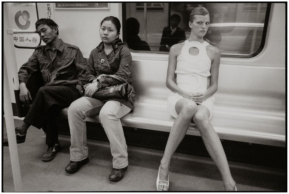 Black and white photograph of three passengers sitting on a subway train, contrasting casual and formal attire.