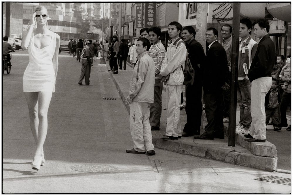 Black and white film strip showing a person in white dress walking down a street while others look on from the sidewalk.