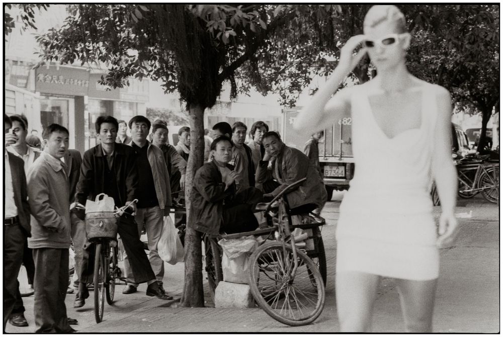 Vintage black and white photograph showing a crowd gathered on a street with bicycles and a person in white clothing.