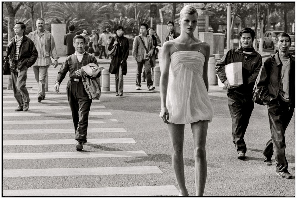 Pedestrians crossing a busy city intersection with crosswalk lines visible in black and white photography.