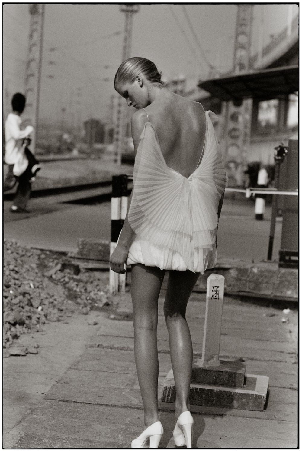 A vintage black and white photograph shows a ballet dancer in a pleated white dress with an open back on a bridge.