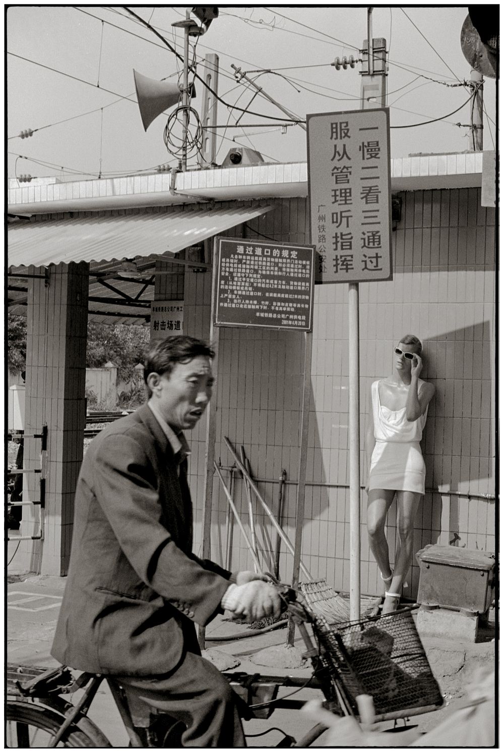 Black and white street photograph from Asia showing a person on a bicycle passing by while another leans against a wall.