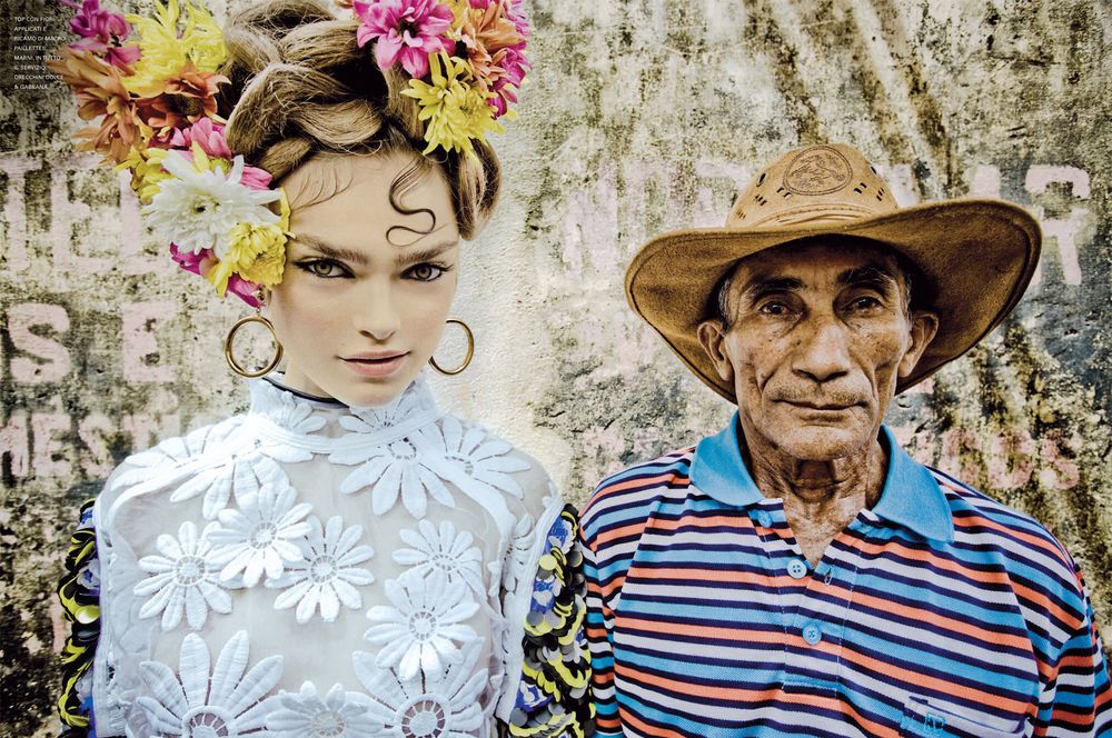 A person wearing a floral white lace dress poses with someone in a striped shirt against a weathered wall.