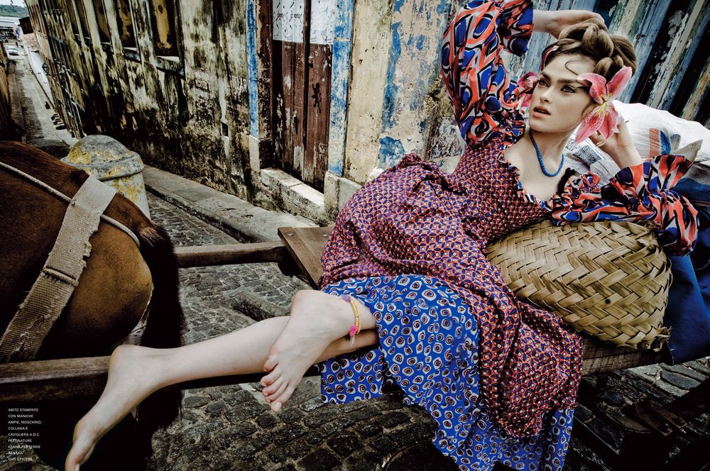 A person in a vintage polka dot dress sits among rustic wooden structures and debris in an outdoor setting.