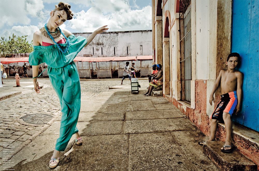 A dancer in a teal dress performs graceful movements in an urban setting with rustic walls and concrete floors.
