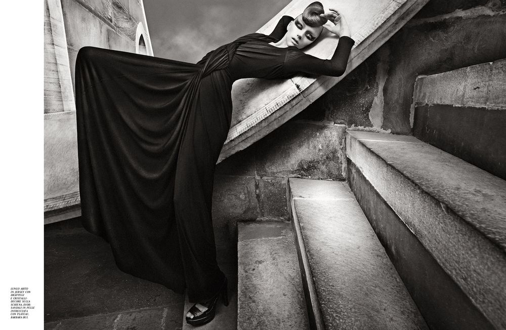 A dramatic black and white photograph shows a figure in flowing black fabric posed elegantly on stone steps.