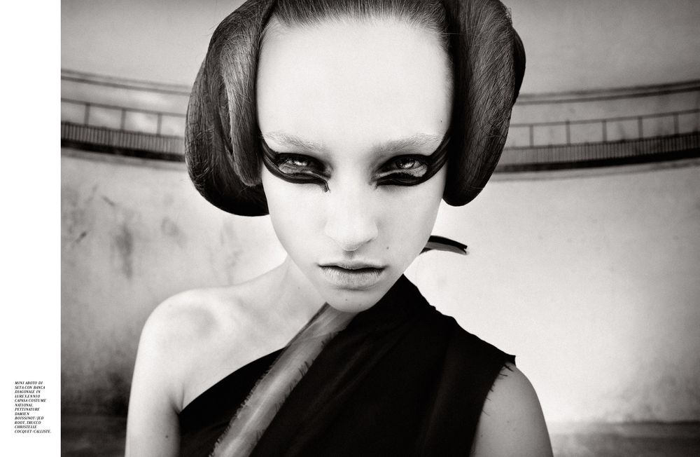 Black and white fashion photograph with dramatic avant-garde makeup and sculptural hairstyle in a minimalist setting.