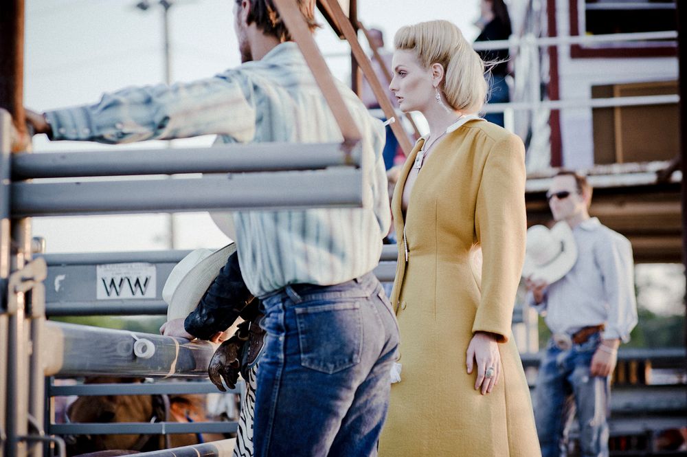 Person in yellow dress stands near a truck at what appears to be a rodeo or western event in vintage style photo.