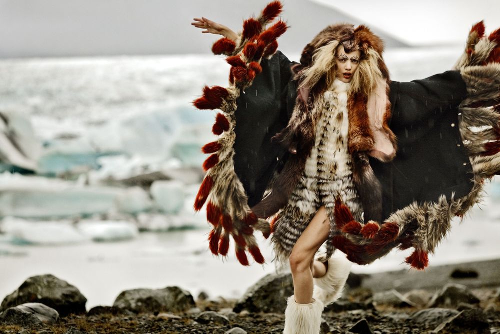 Fashion model in dark feathered costume poses dramatically against rocky coastal landscape.