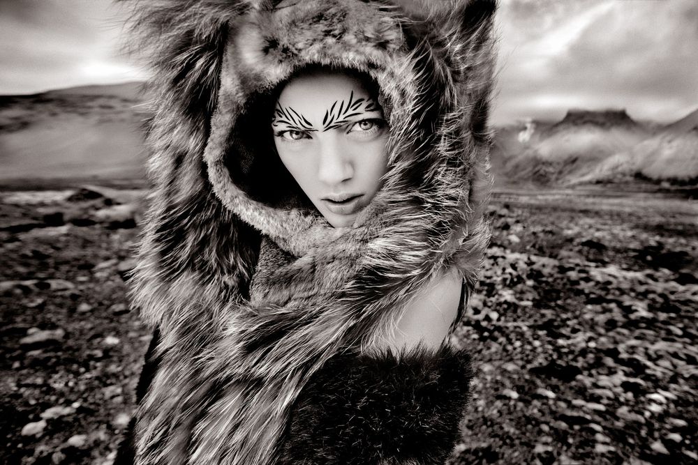 Black and white artistic portrait with fur hood against dramatic landscape background.