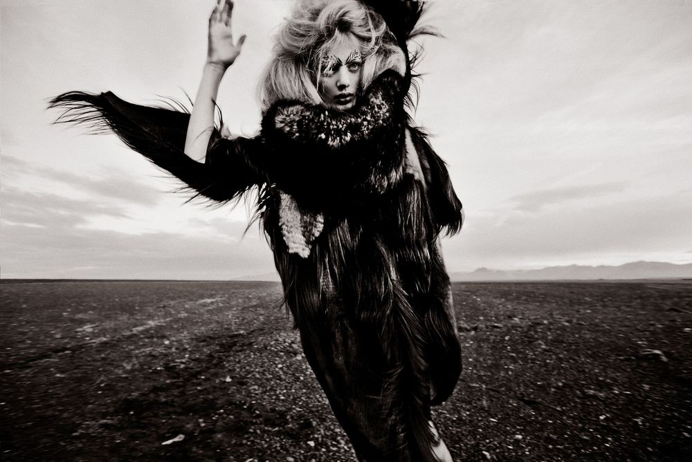 A figure in a flowing black dress and feathered collar dances expressively across a stark outdoor landscape.
