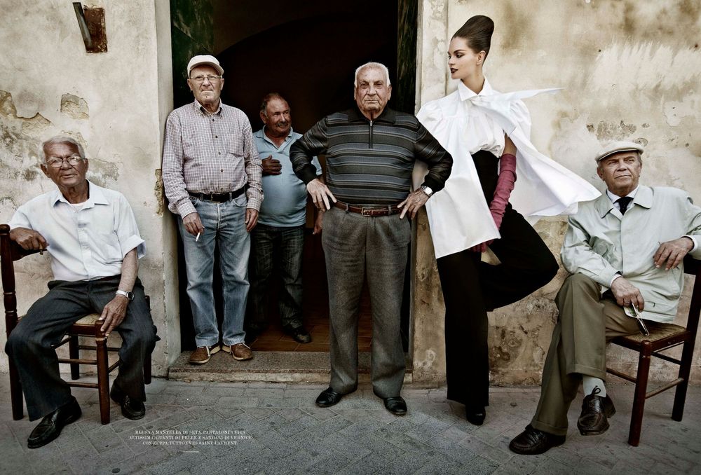 A row of elderly men sit in chairs along a white stone wall in a Mediterranean village setting.