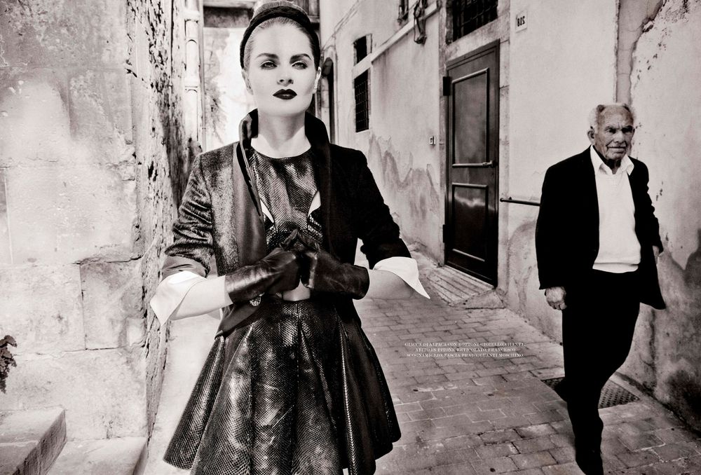 Black and white street scene showing a person in a dark dress on a narrow European alley with someone walking behind.