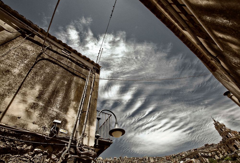 Dramatic rippled clouds sweep across a moody sky above weathered building walls in an architectural HDR photograph.