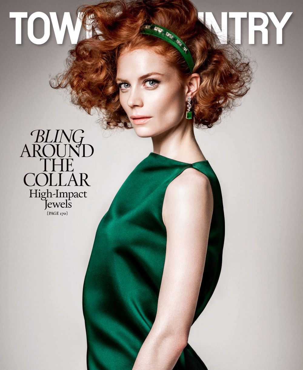 Fashion model in emerald green sleeveless dress with red curly hair and headband poses against neutral background.