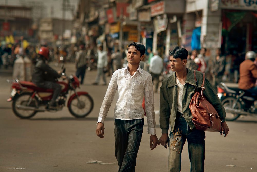 Two friends walk down a busy street with motorcycles and shops in the background.