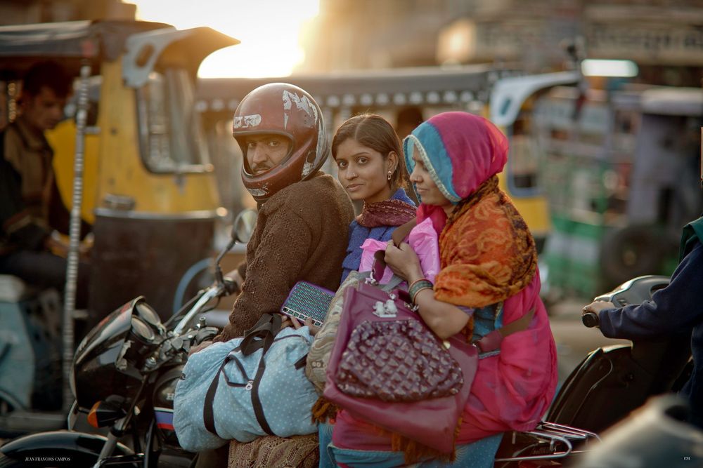 A person rides a motorcycle with passengers through a busy city street during daytime.