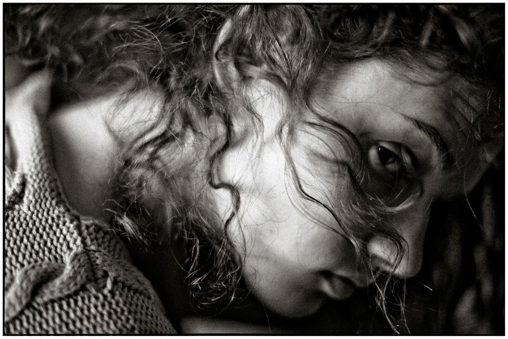 Black and white close-up portrait of a young child with curly hair and an intense gaze looking directly at camera.