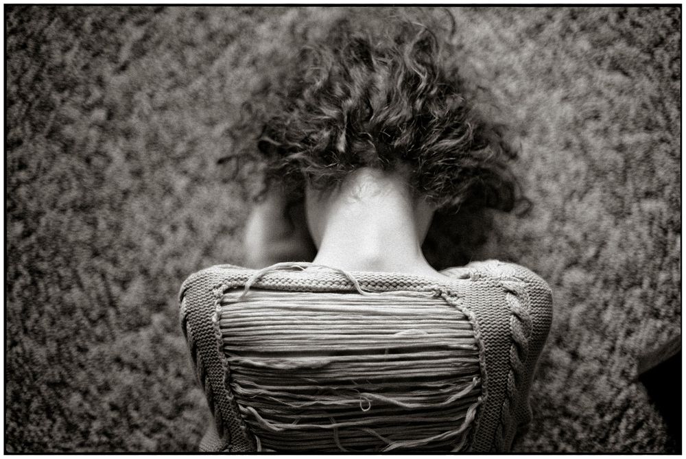 Black and white photo of a person curled over a backpack, dark curly hair covering their face.