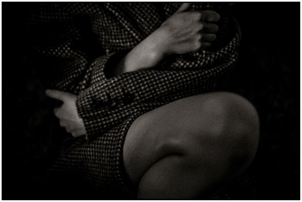 Black and white close-up of hands clasping a knee in a tweed jacket, moody dark background.