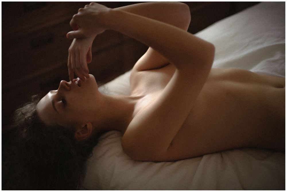 Woman lying on bed in natural light with arms raised above head in relaxed pose.