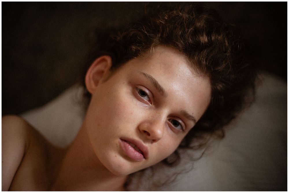Portrait of a woman with curly hair resting on white pillows, looking directly at camera with natural makeup and bare shoulders.
