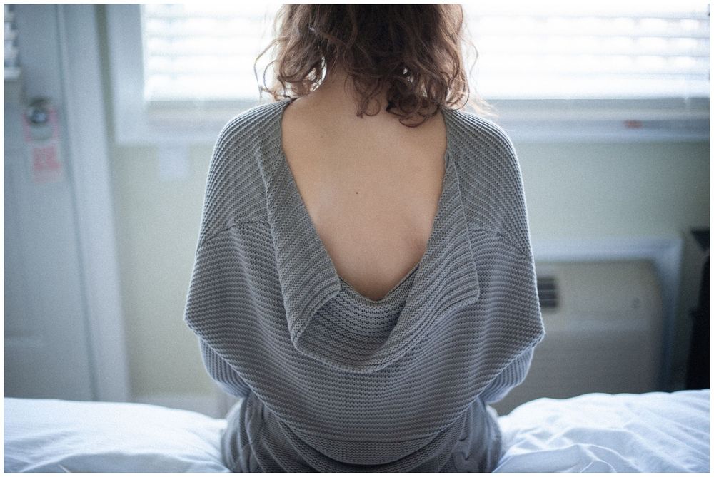 Woman in striped sweater sitting on bed with back turned showing draped neckline in bright room.