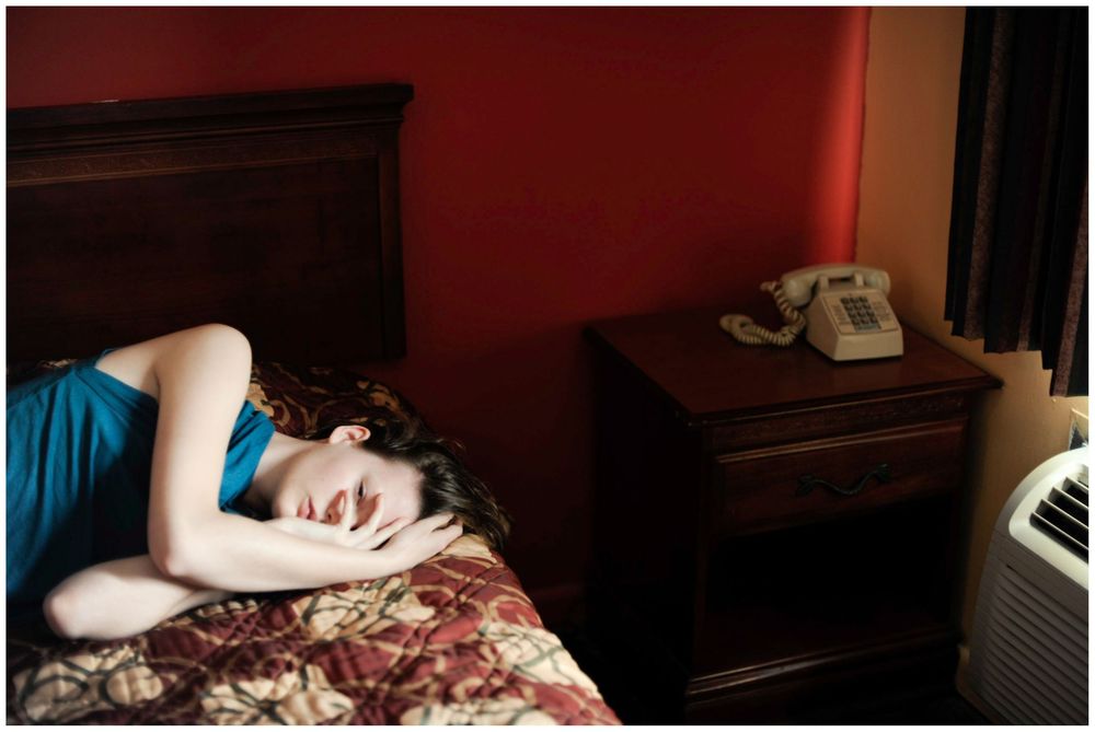 Woman in blue dress sleeping on a floral bedspread in a motel room with red walls and vintage phone.