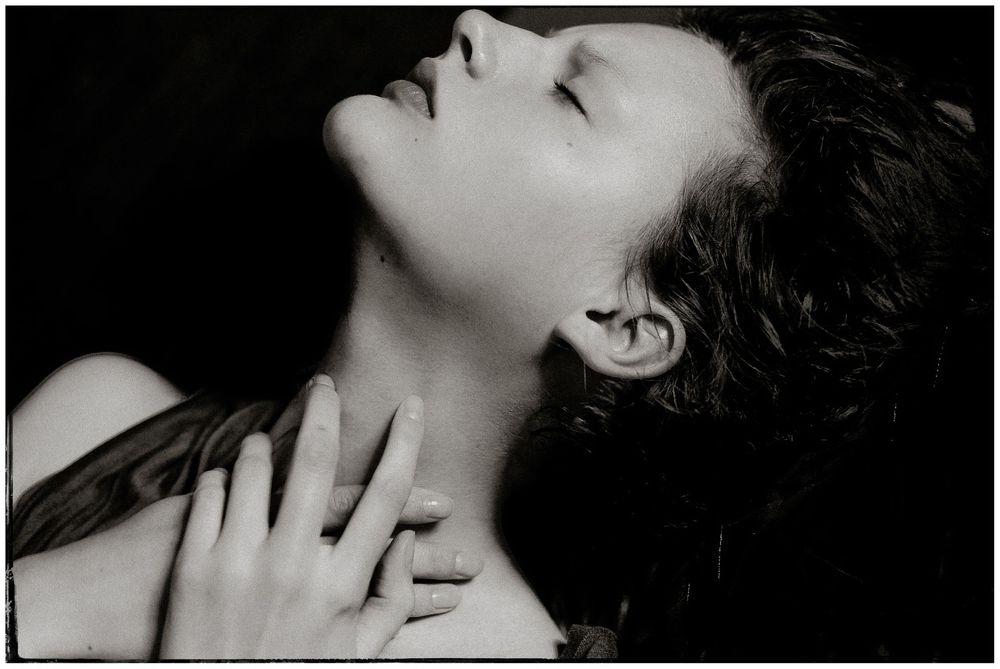 Black and white portrait of a woman with dark hair tilted back, hand on chest, artistic photography.