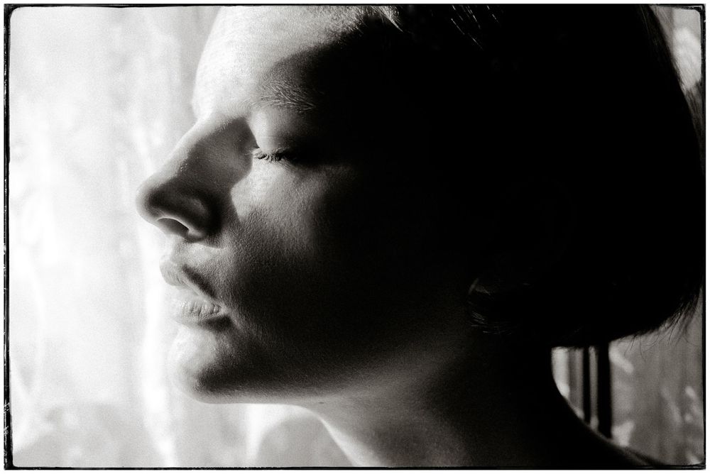 Black and white profile portrait of a woman with closed eyes, soft light illuminating her face near a window.