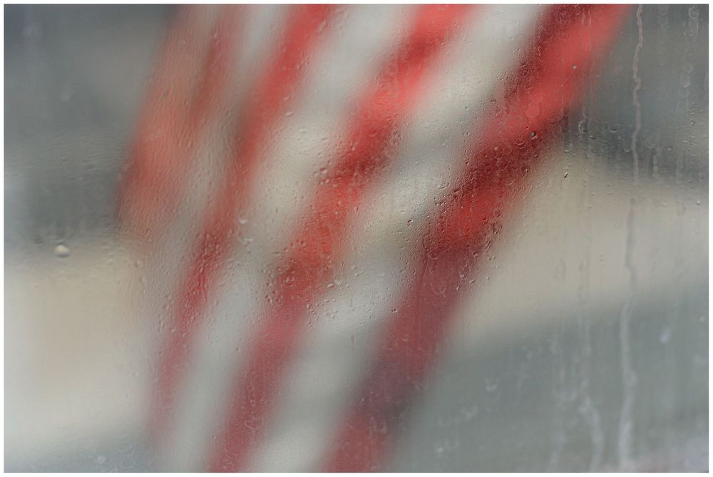 Blurred abstract image showing red and white diagonal stripes through a textured, rain-streaked or frosted glass surface.
