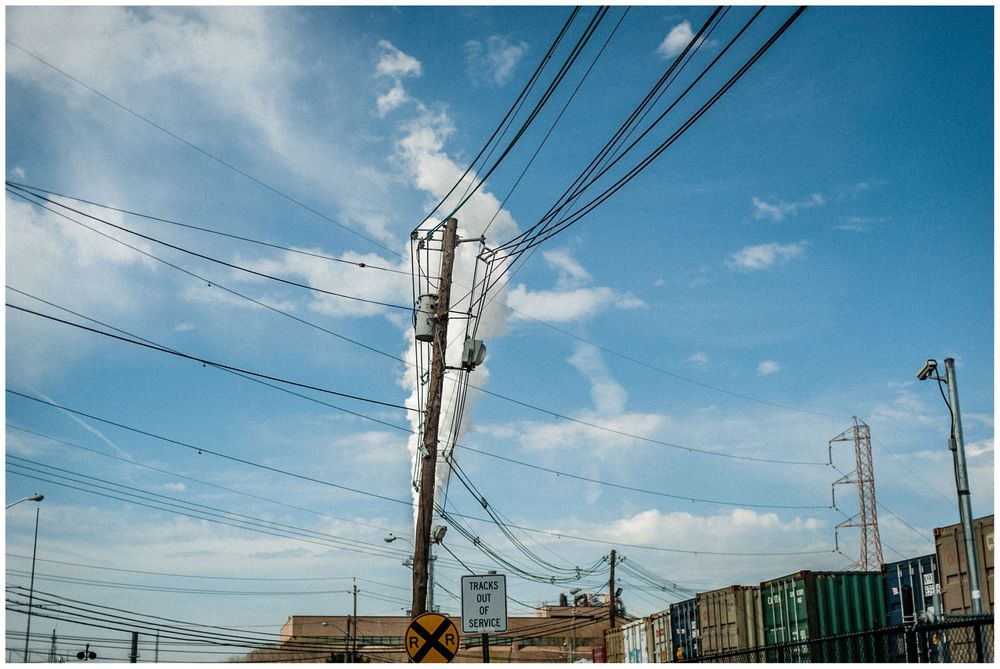 Utility pole with power lines against blue sky with clouds and industrial buildings in background.