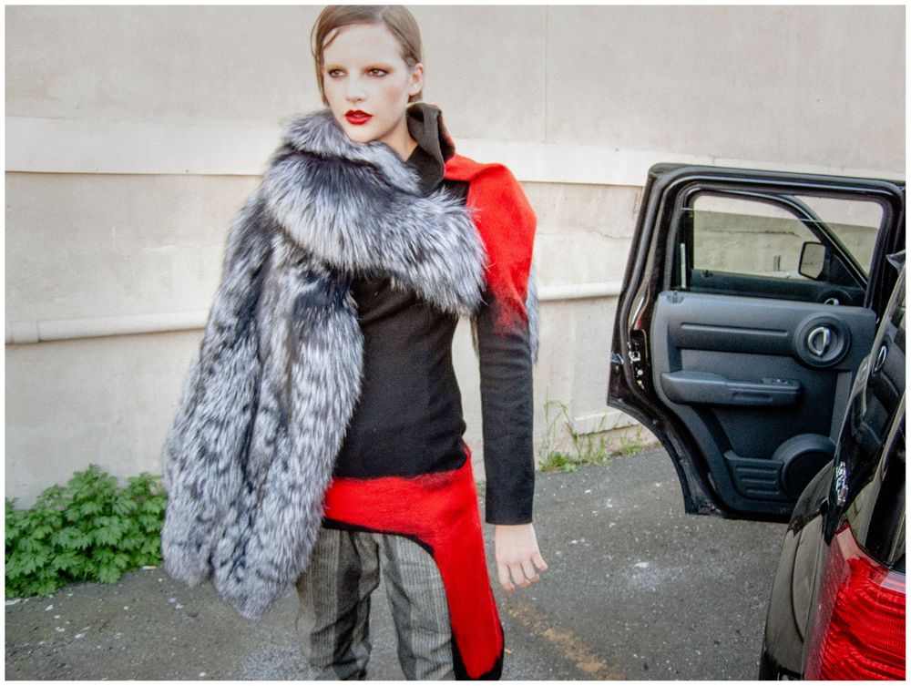 Woman in black outfit with red accents and silver fur coat posing next to open car door.