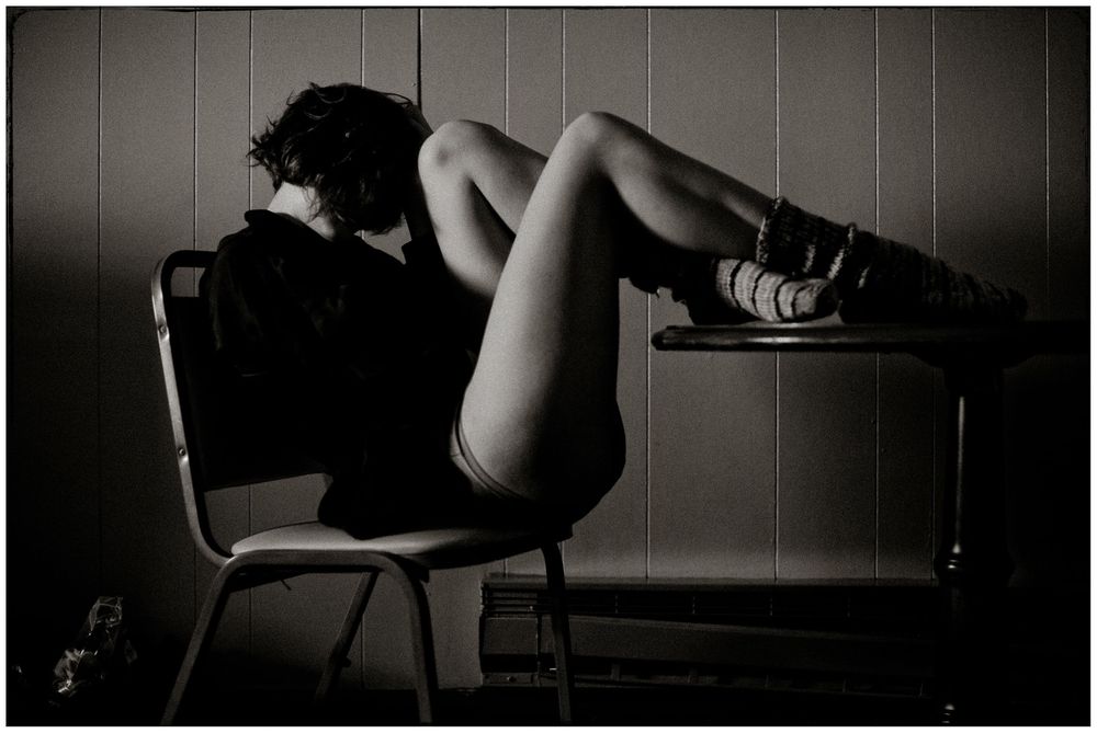Black and white photo of a woman sitting on a chair with legs extended, wearing sneakers in an artistic pose.