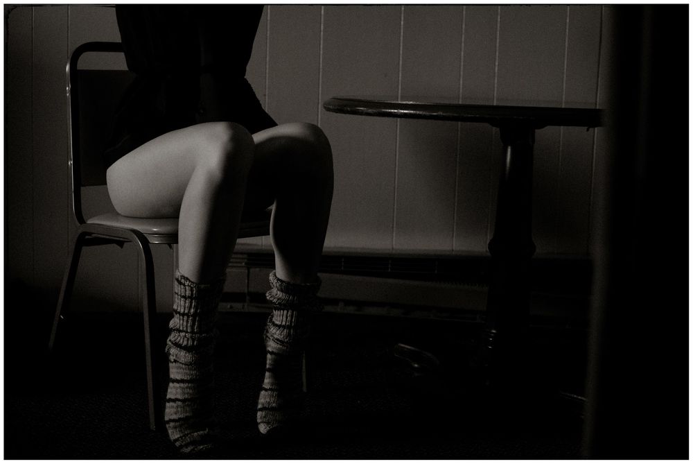 Black and white artistic photograph of a woman sitting on a chair in a dimly lit room with minimalist furniture.